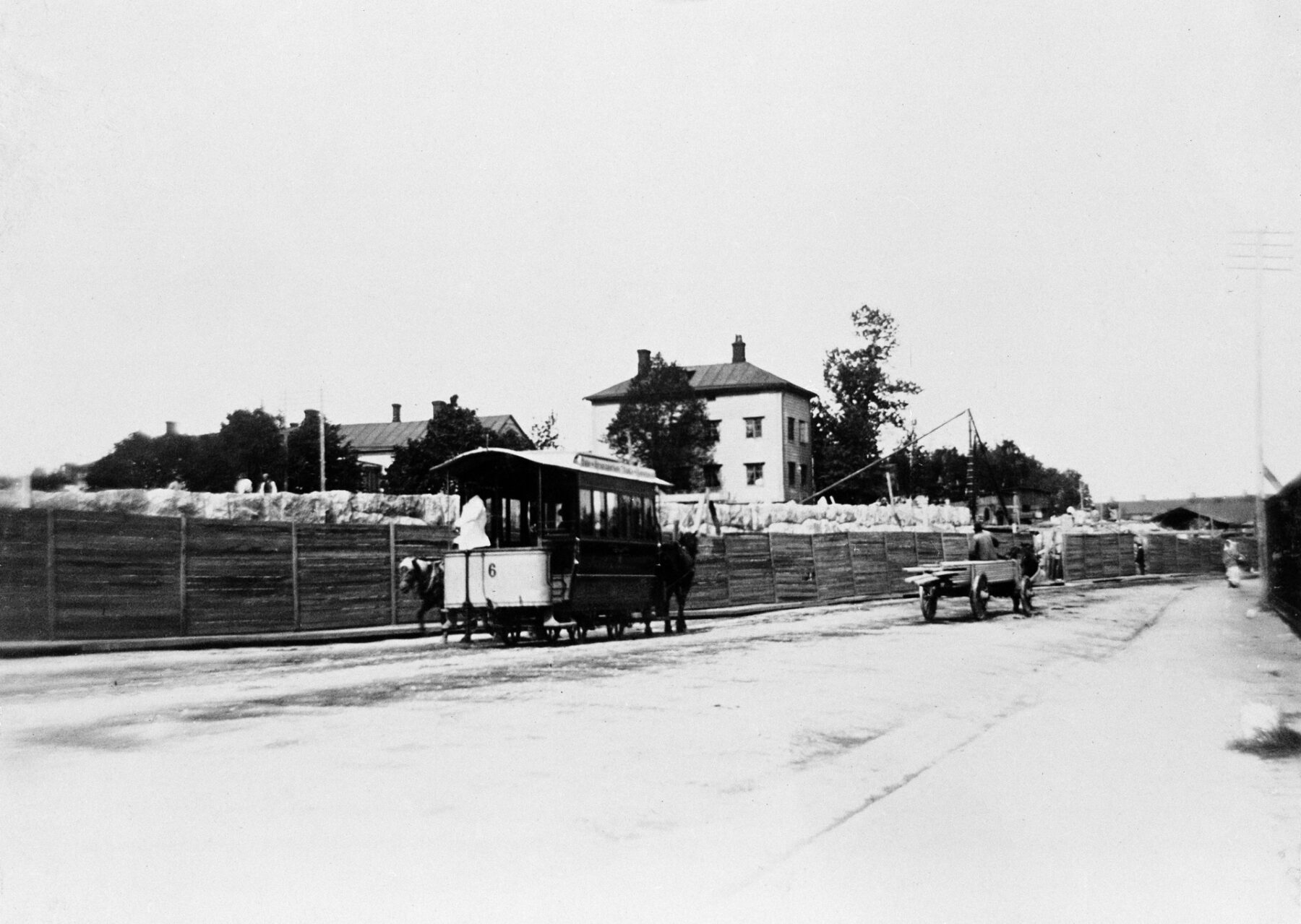 A horse-drawn tram sits on a road next to a wooden fence, with buildings visible in the background.