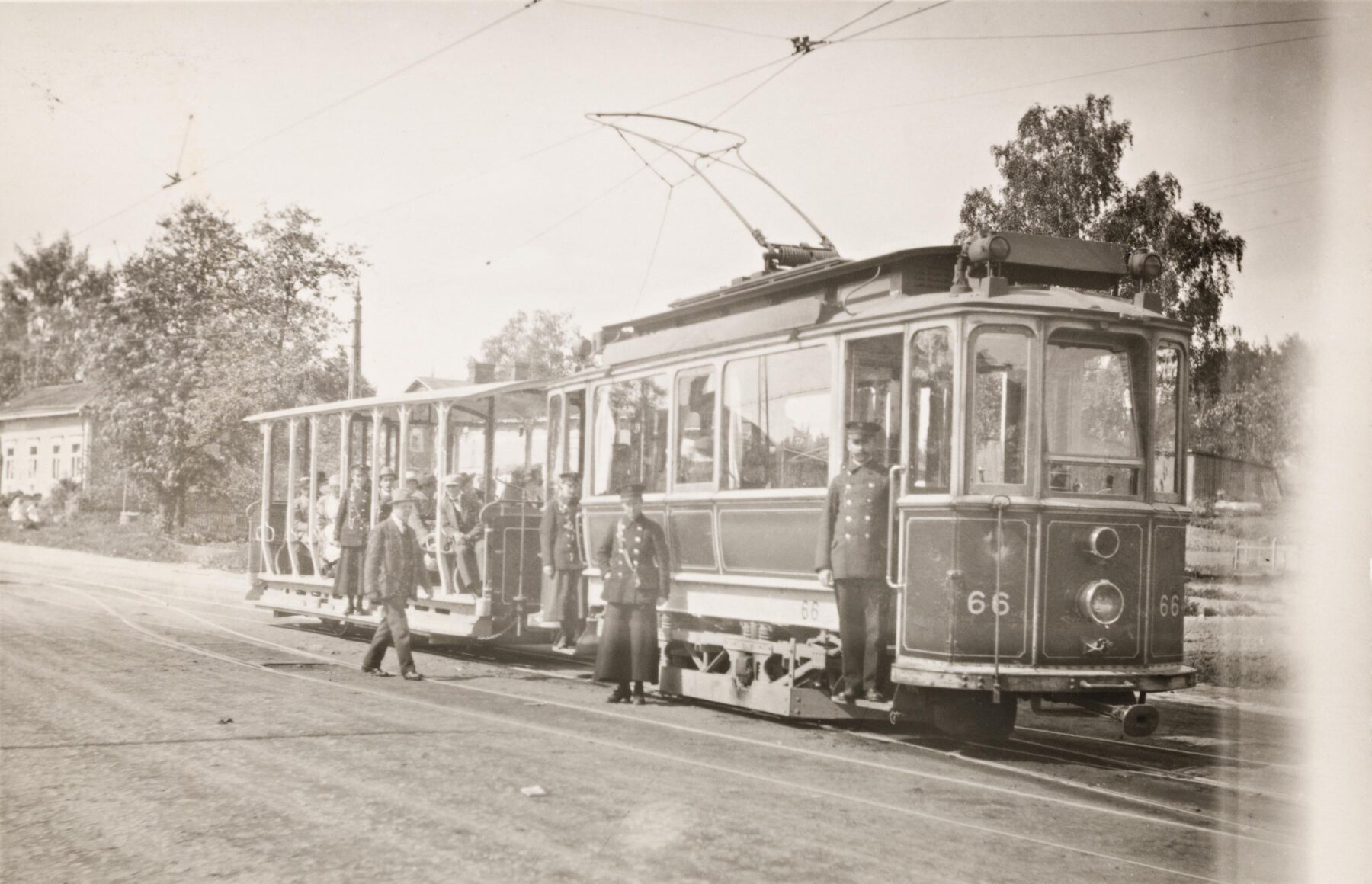 An old tram car towing an open-air trailer full of passengers stands idle on tracks while people in uniforms stand nearby, in a vintage black and white photograph.
