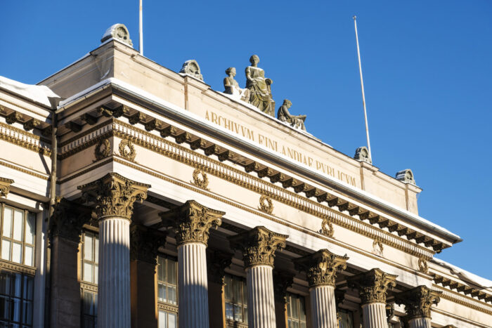 Several statues are visible above the pillars that decorate the facade of a building where letters spell “National Archives of Finland” in Latin.