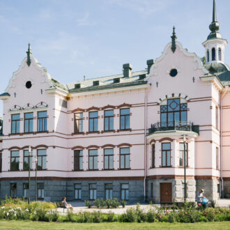 A 19th-century building with a granite foundation and pink stucco walls stands against a blue sky surrounded by trees and grass.