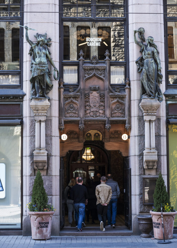 Two statues stand atop the columns that flank a doorway on a main street with people and trams passing by.
