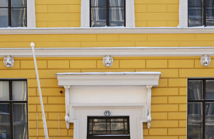 On a yellow two-storey building, small lion’s-head sculptures are visible at regular intervals above the ground-floor windows.