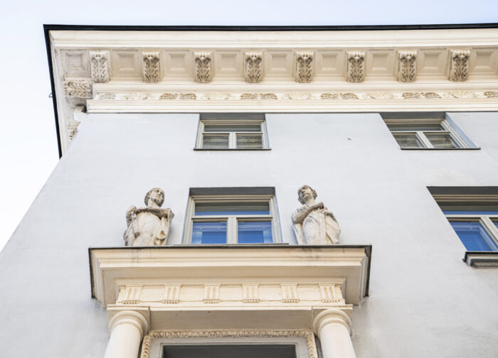 Four statues flank the third-floor windows on the corner of an old-fashioned light-blue building.
