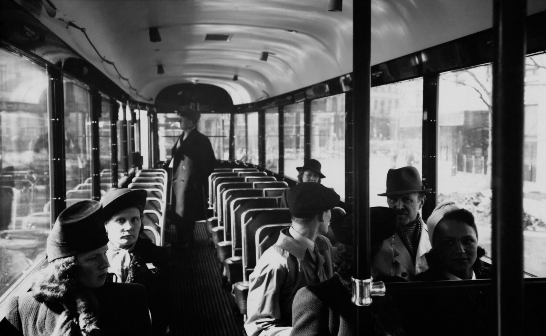 People in 1940s clothing sit on a tram with large windows and rows of empty seats with one man standing at the back.
