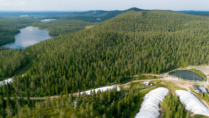An aerial shot shows a tree-covered landscape, a lake and several white covers running down ski slopes toward a small pond.