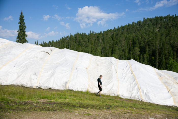 A person walks past a large snow mound covered in white sheeting on a grassy slope with a forested hill in the background.