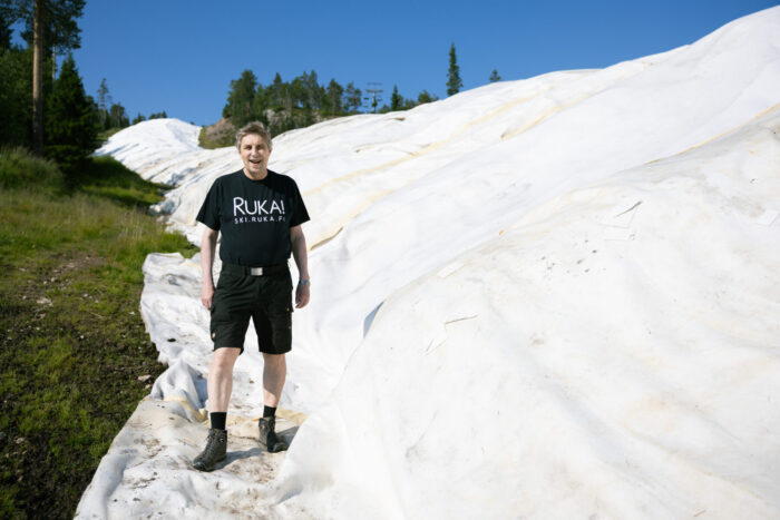  A person in a T-shirt and shorts stands on a white tarp with green grass and trees visible behind them and a clear blue sky above.