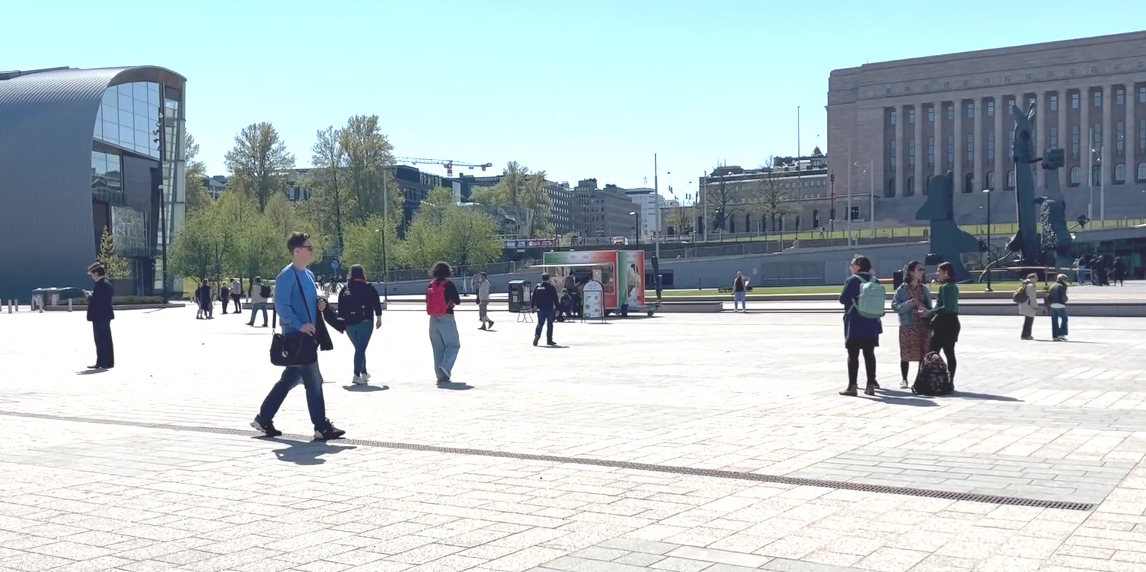 En un día soleado, varias personas caminan en diferentes direcciones por una plaza rodeada de edificios monumentales.