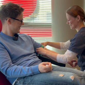 A man is sitting in a chair beside a rack of medical equipment while a woman rolls up his sleeve in preparation for him to donate blood.