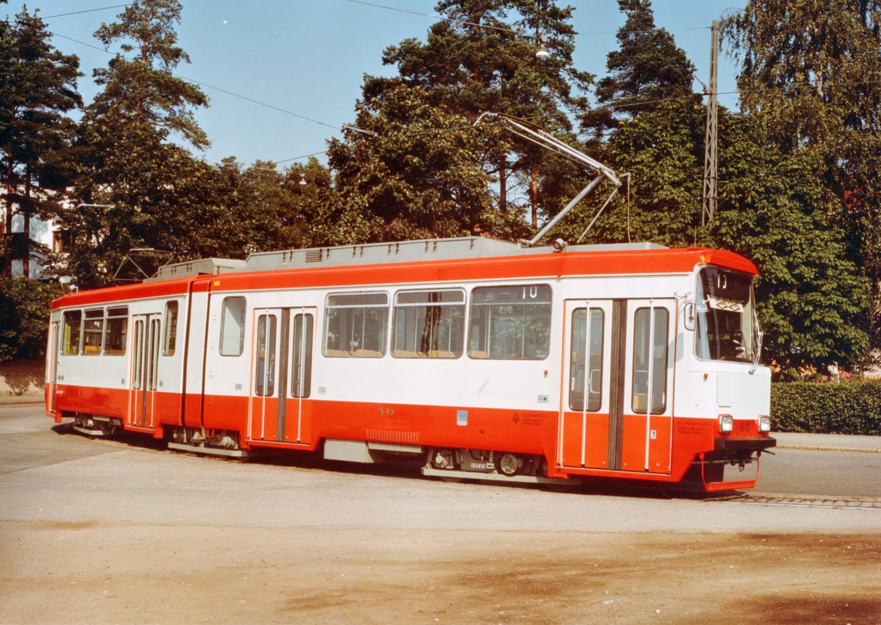 An orange and grey tram sits on a rail with greenery and tall trees in the background under a clear, bright sky.