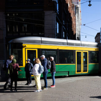 Bajo un cielo depejado, en un escenario urbano, un tranvía verde y amarillo circula por las vías y pasa junto a un grupo de peatones.