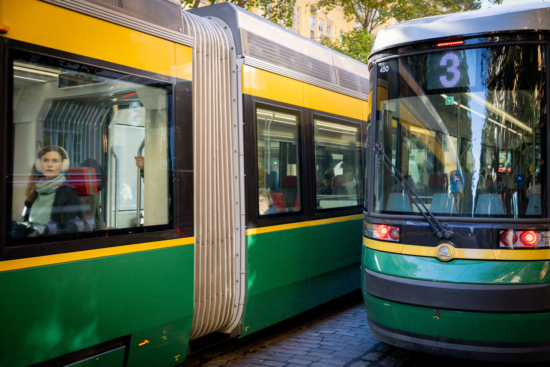 Two green and yellow trams pass each other on a city street, with passengers visible through the windows.