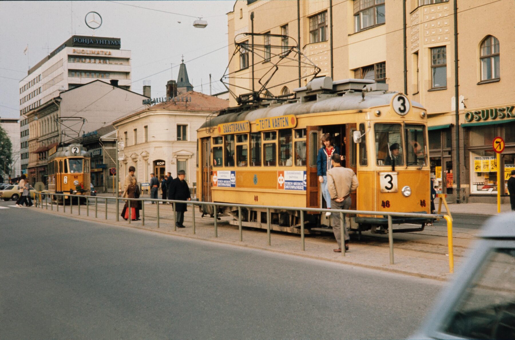 A yellow tram sits on tracks in a 1970s urban setting, with people boarding and walking nearby.