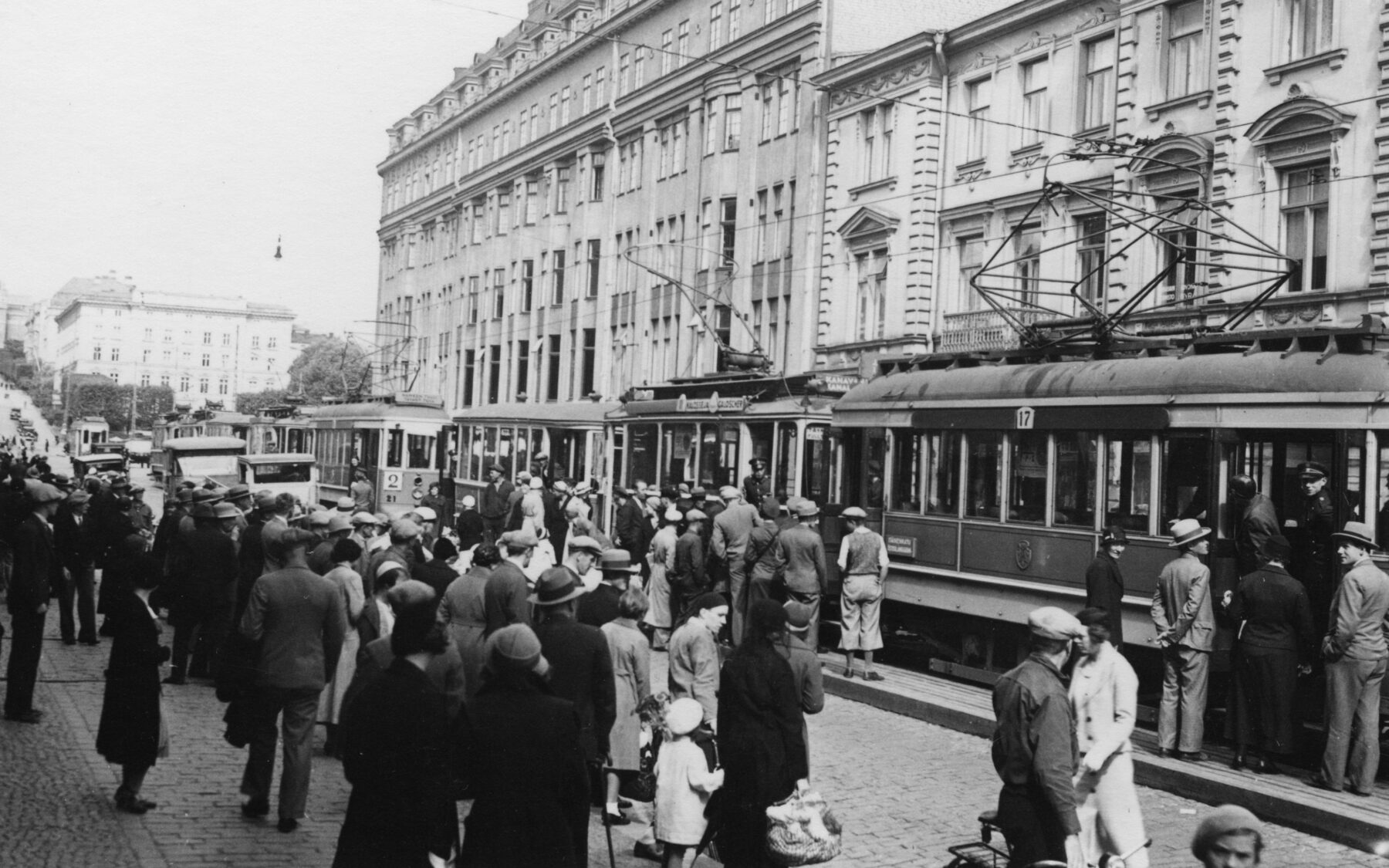 A crowd gathers near a line of trams on a busy city street in the 1930s.