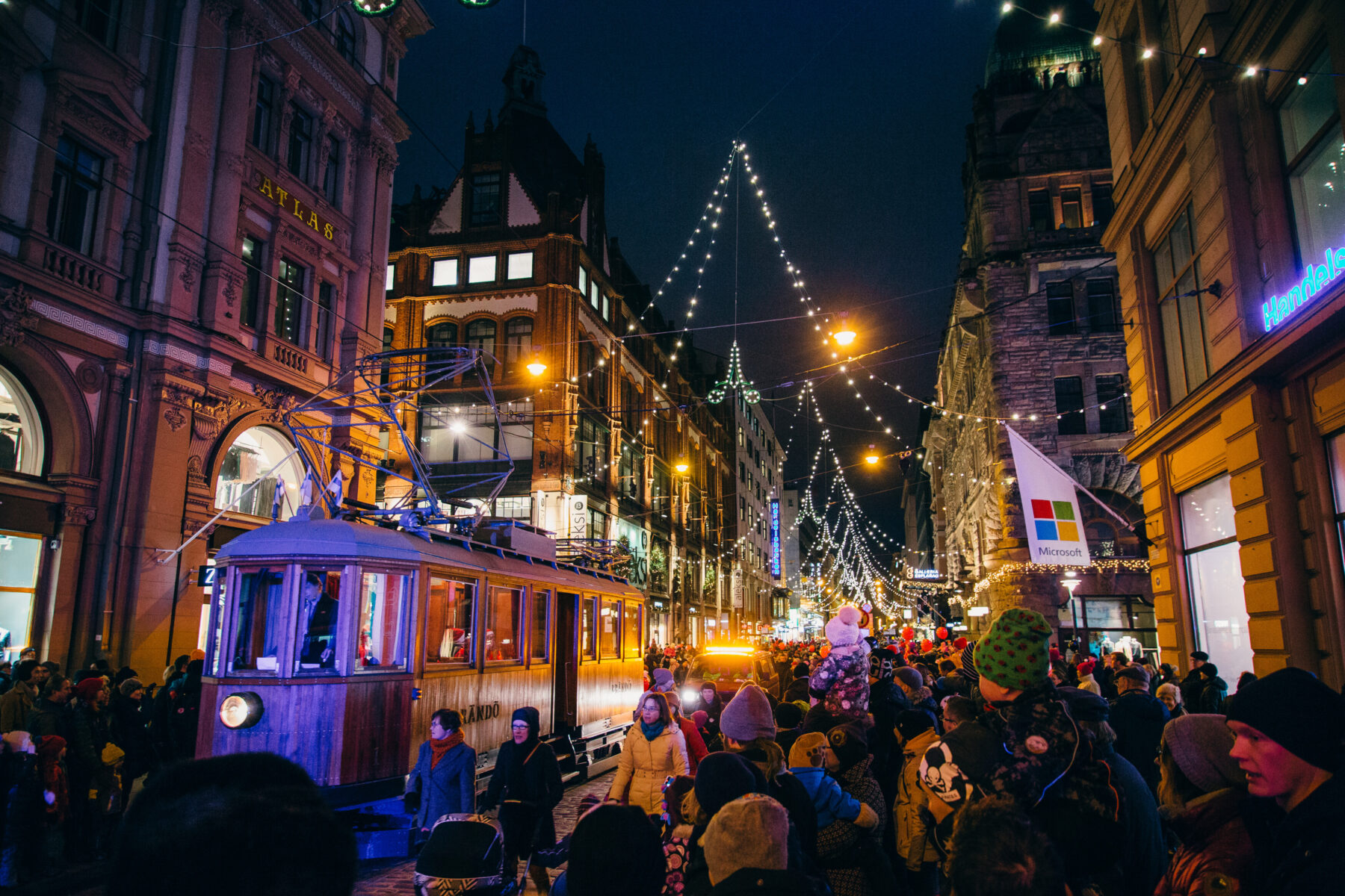 Crowds gather at night along a street lined with tall buildings and sparkling Christmas lights as an old-fashioned tram glides by.