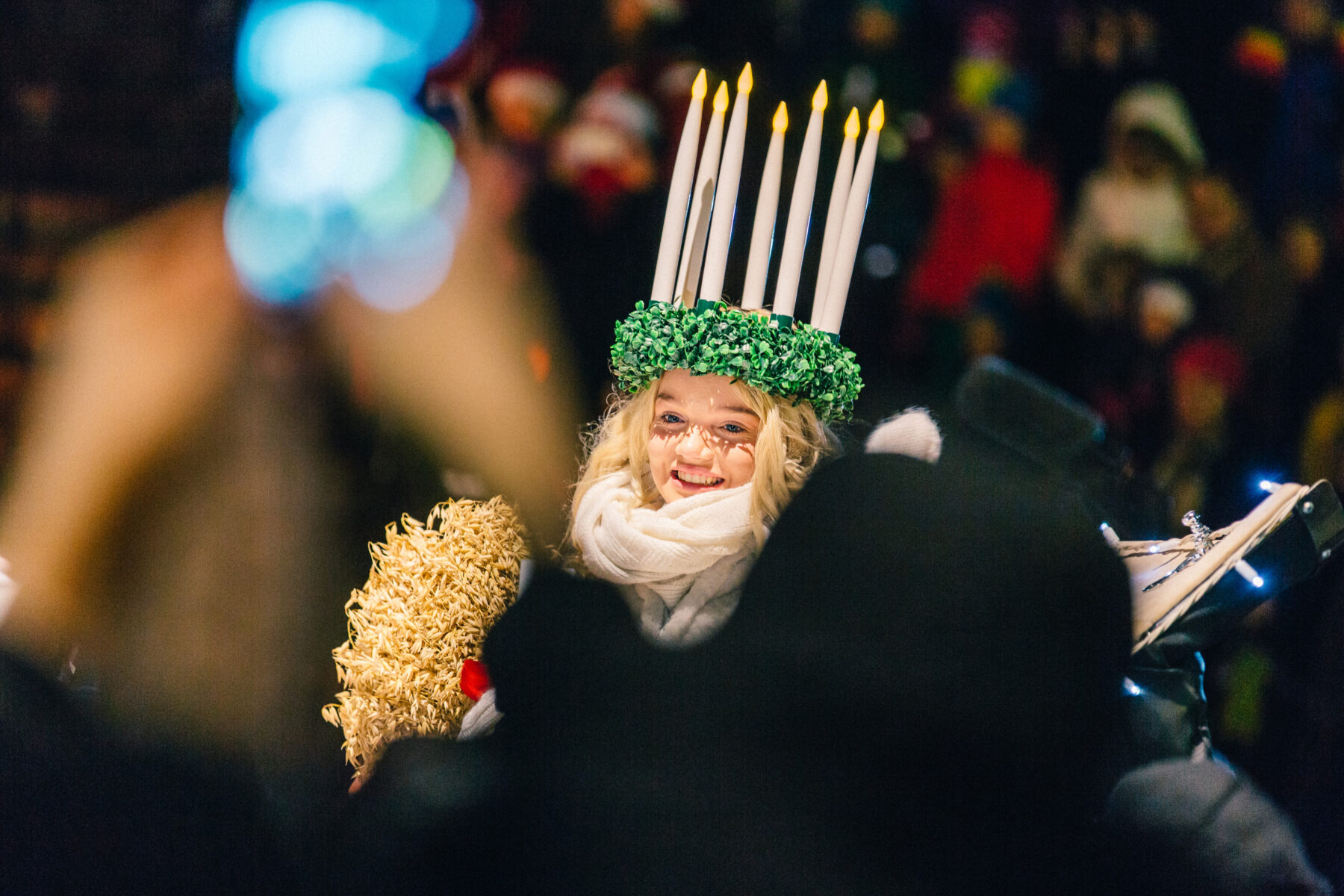 A smiling woman in white portrays Saint Lucia, wearing a crown of candles and waving to a crowd of onlookers.