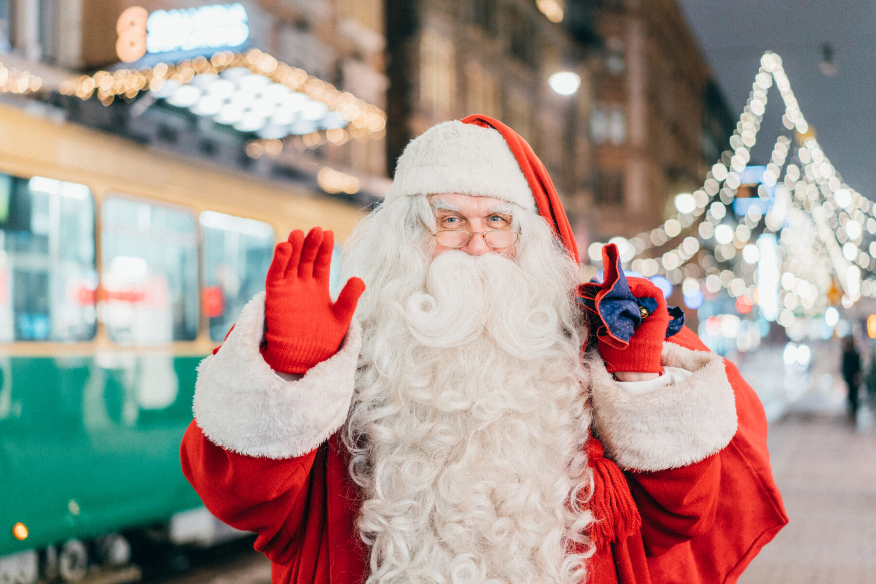 Santa Claus, with a long white beard and red gloves, smiles and waves, carrying a red sack of gifts along a downtown street lit by Christmas lights.