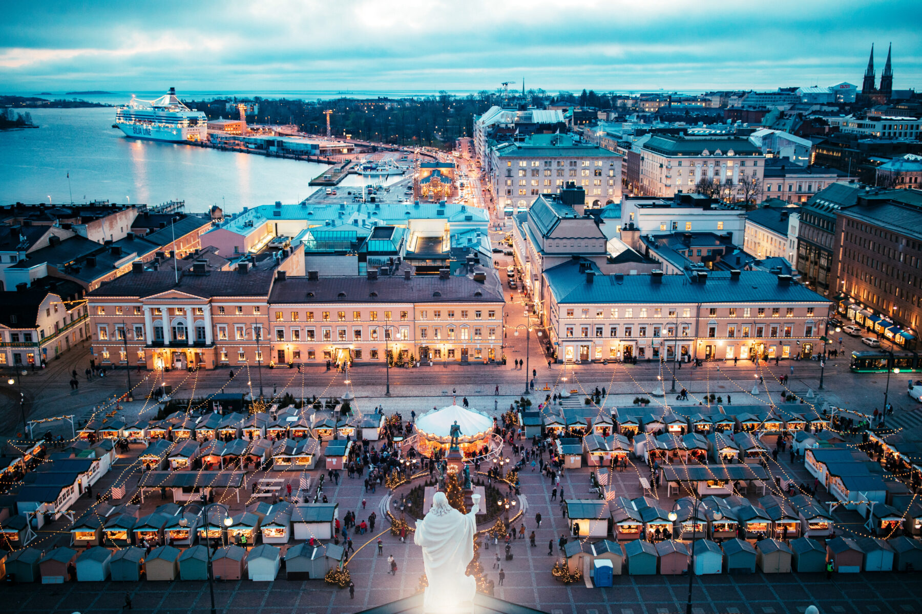 From a high viewpoint, warmly lit stalls are visible in a city square, while farther away a calm harbour is illuminated by soft twilight.