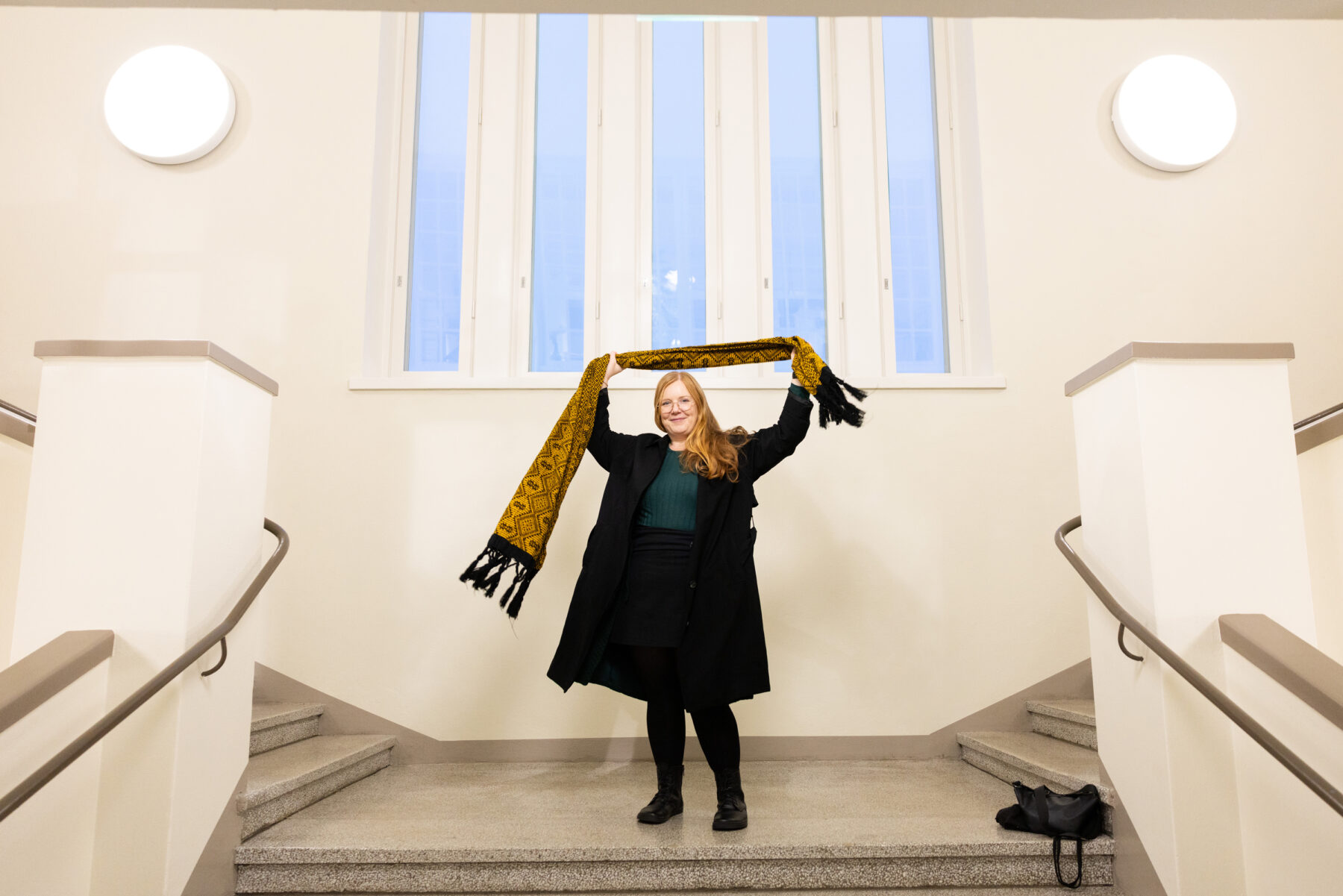 A woman smiles and holds a scarf above her head while standing on the landing of a wide double staircase.