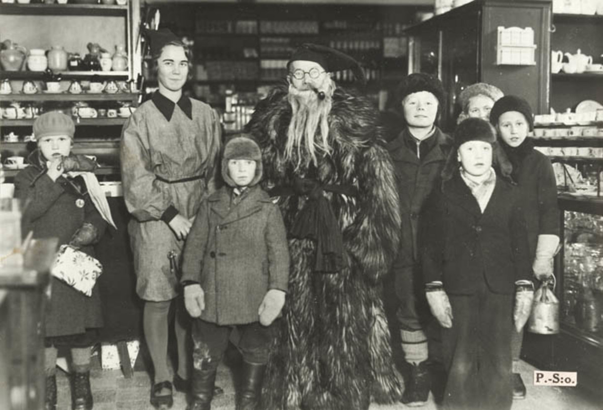 Father Christmas in a dark fur coat holds a pipe while standing beside a woman dressed as an elf in a shop in a 1930s black-and-white photo.