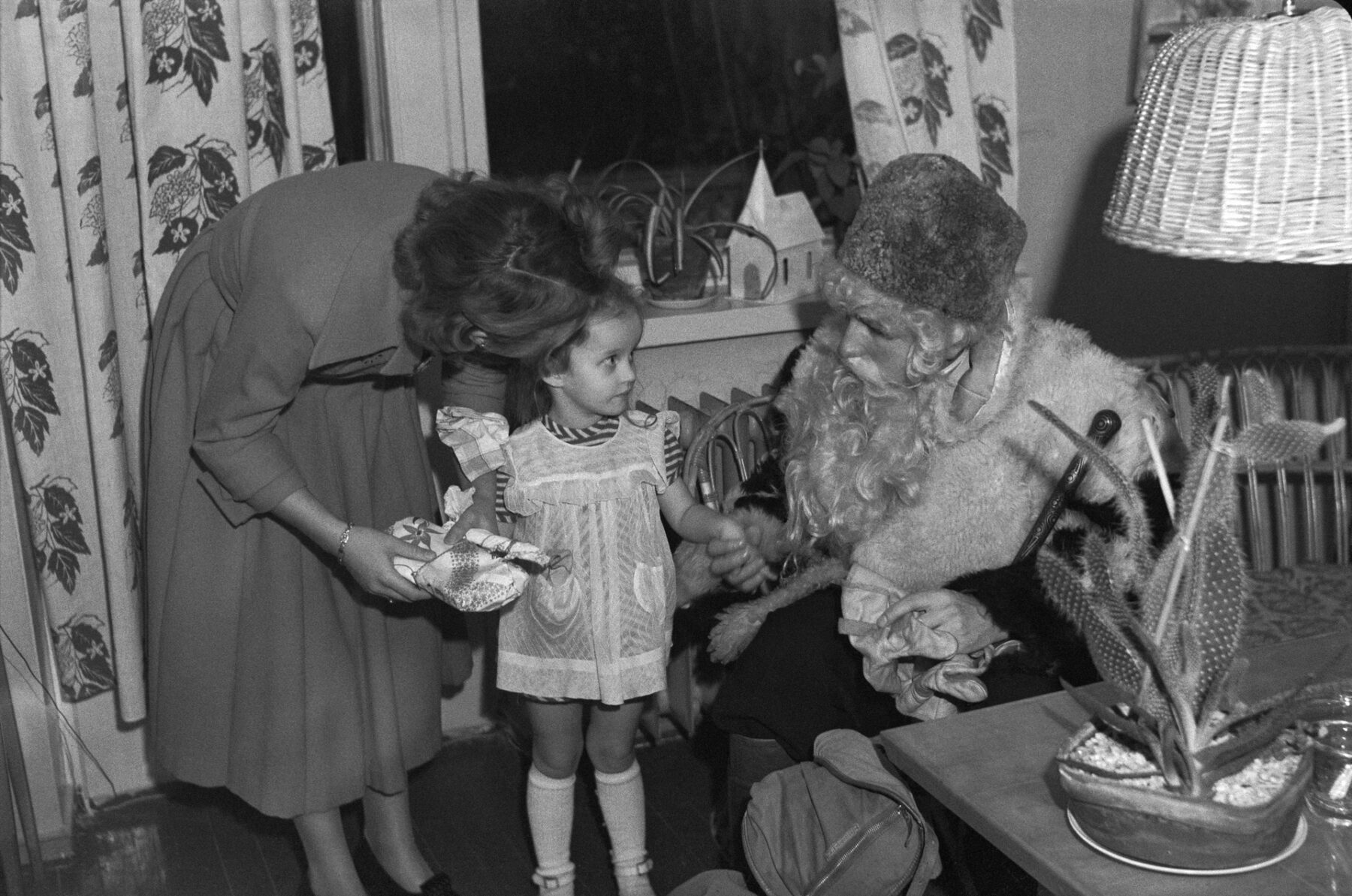 A young girl in the 1950s looks at a man dressed as Santa in her family’s living room as her mother bends down to speak encouragingly to her.