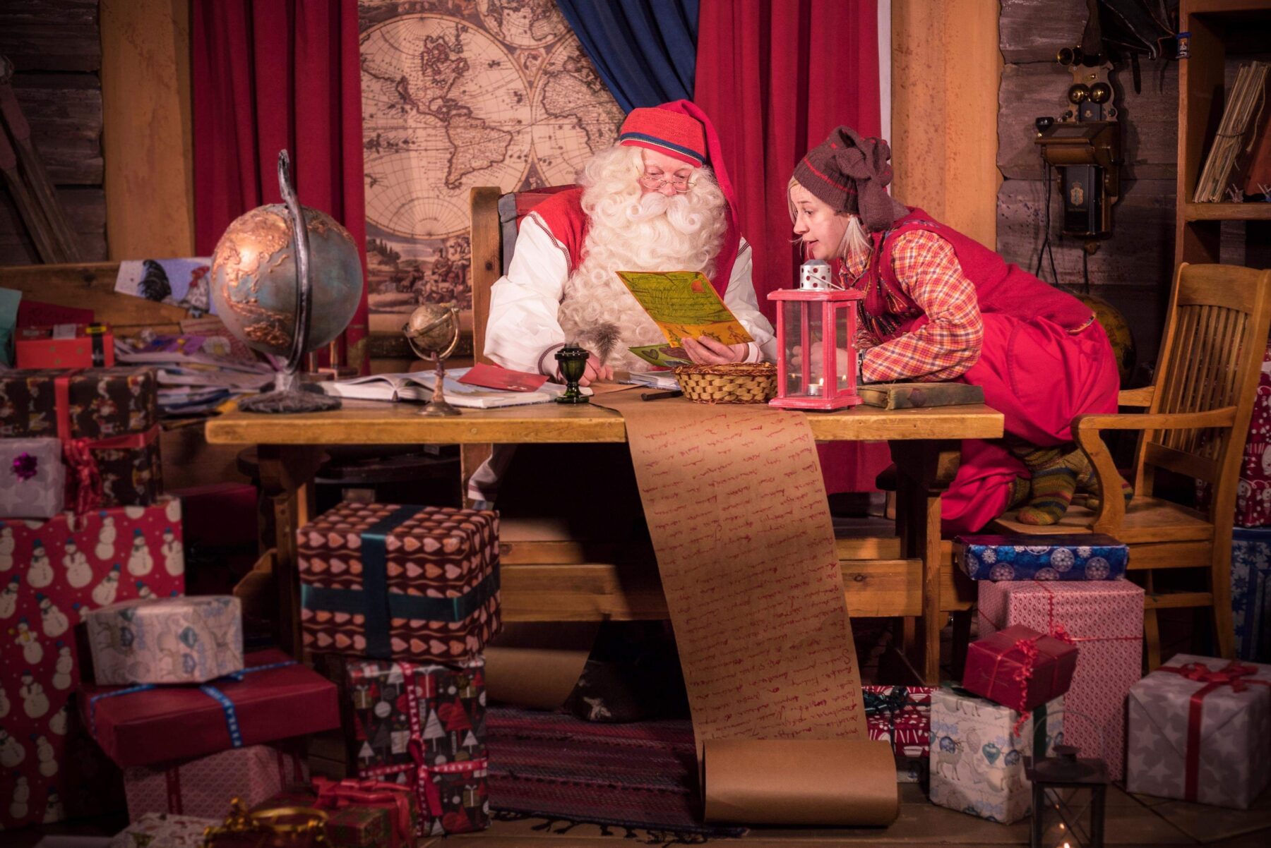 Santa shows an elf a card as they sit at a wooden desk surrounded by presents with a long parchment scroll hanging to the floor.