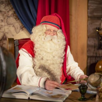 Papá Noel, con un gorro rojo y su barba blanca, posa en su cabaña, sentado ante un escritorio de madera y escribiendo con una pluma de ave.