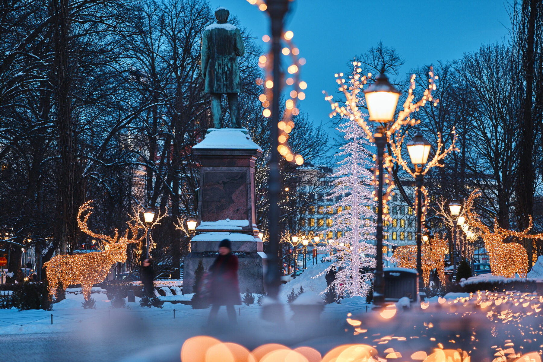 Christmas lights and old streetlamps illuminate a snowy park at dusk, with a tall bronze statue covered in snow standing over glowing reindeer light sculptures nearby.