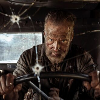 A bearded man, Jorma Tommila as the character Aatami Korpi, is covered in dirt and sweat while driving a vehicle that has several bullet holes in the windshield.