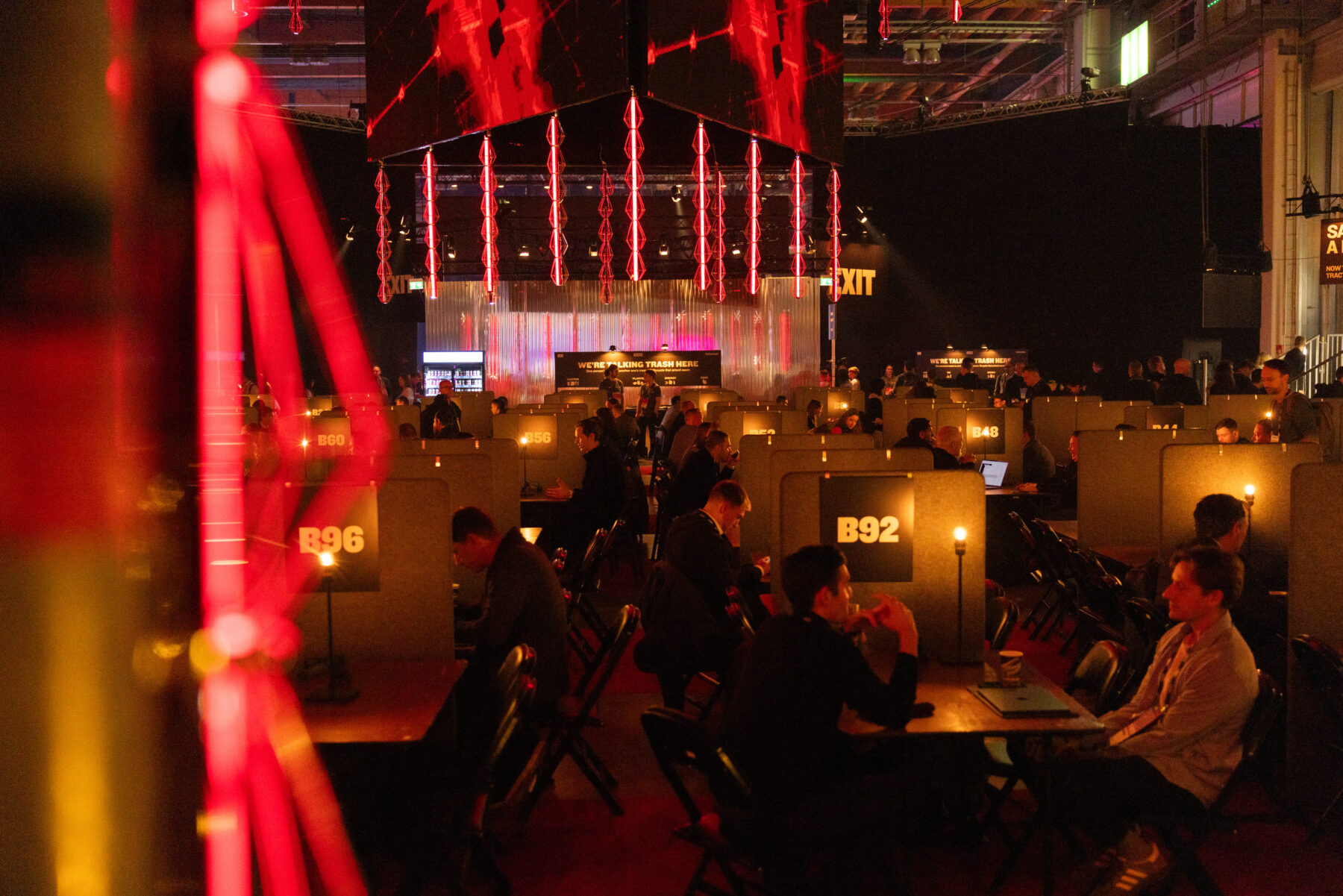 في قاعة مناسبات مظلمة، يجلس العديد من الأشخاص على طاولات مرتبة، بينما تتدلى مصابيح إضاءة حمراء من السقف.