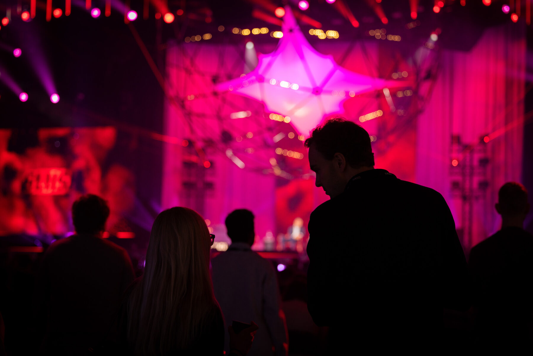 ظلال تواجه مسرحًا مليئًا بالألوان الزاهية والأضواء الوردية الساطعة والزخارف الهندسية.