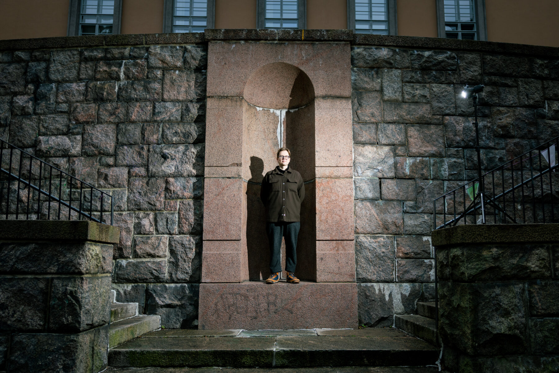 El escritor finlandés Juhani Karila posa en el centro de un nicho de granito rosado, enmarcado por una pared simétrica de piedra gris, con dos tramos de escaleras a ambos lados.