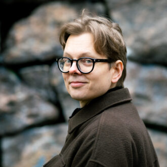 O escritor finlandês Juhani Karila, usando óculos e um casaco marrom, olha por cima do ombro para a câmera, tendo como pano de fundo uma parede de pedra rústica.