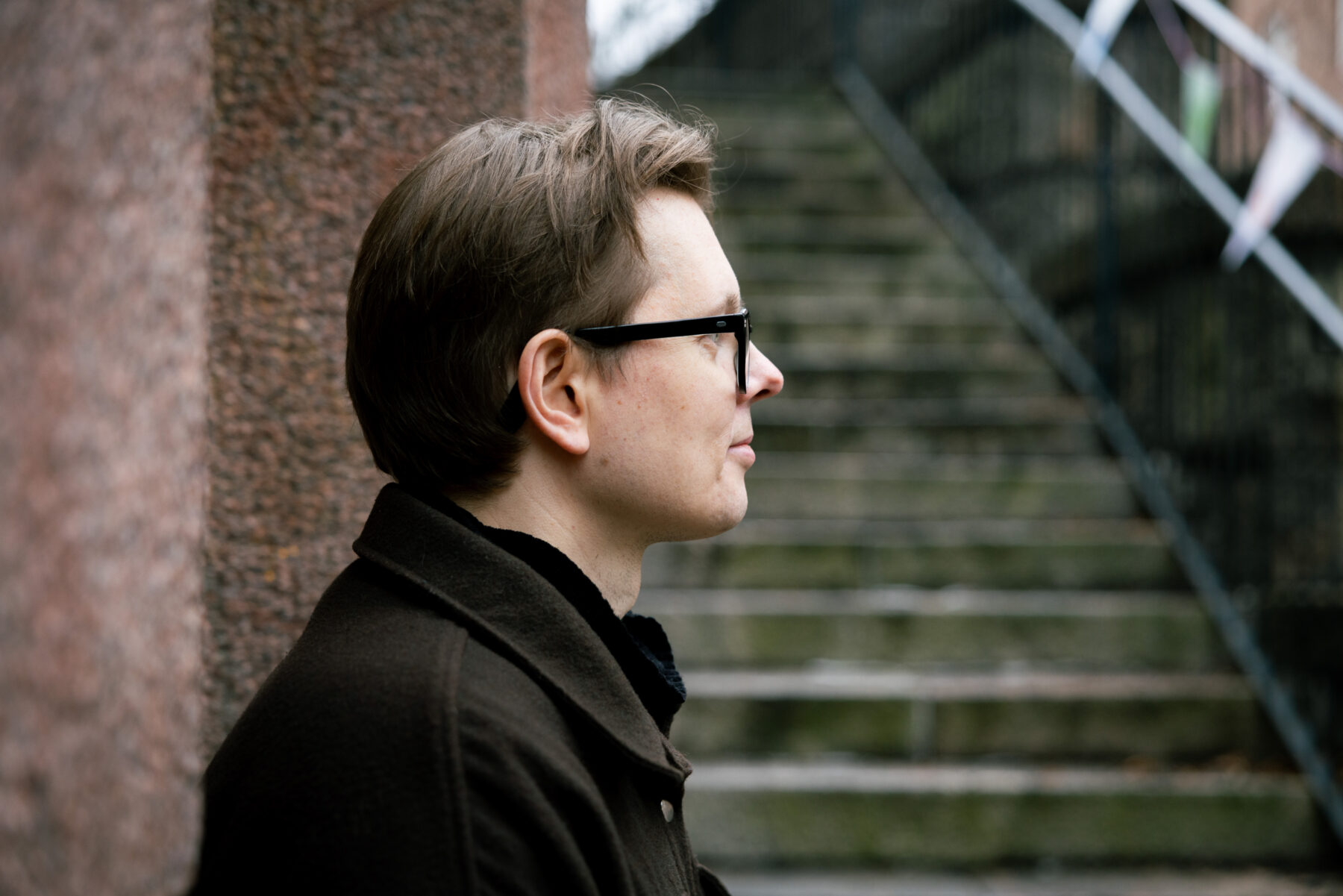 El escritor finlandés Juhani Karila, con gafas y una chaqueta marrón, posa de perfil junto a un muro de granito, mientras que al fondo, detrás de él, se ven unas escaleras.