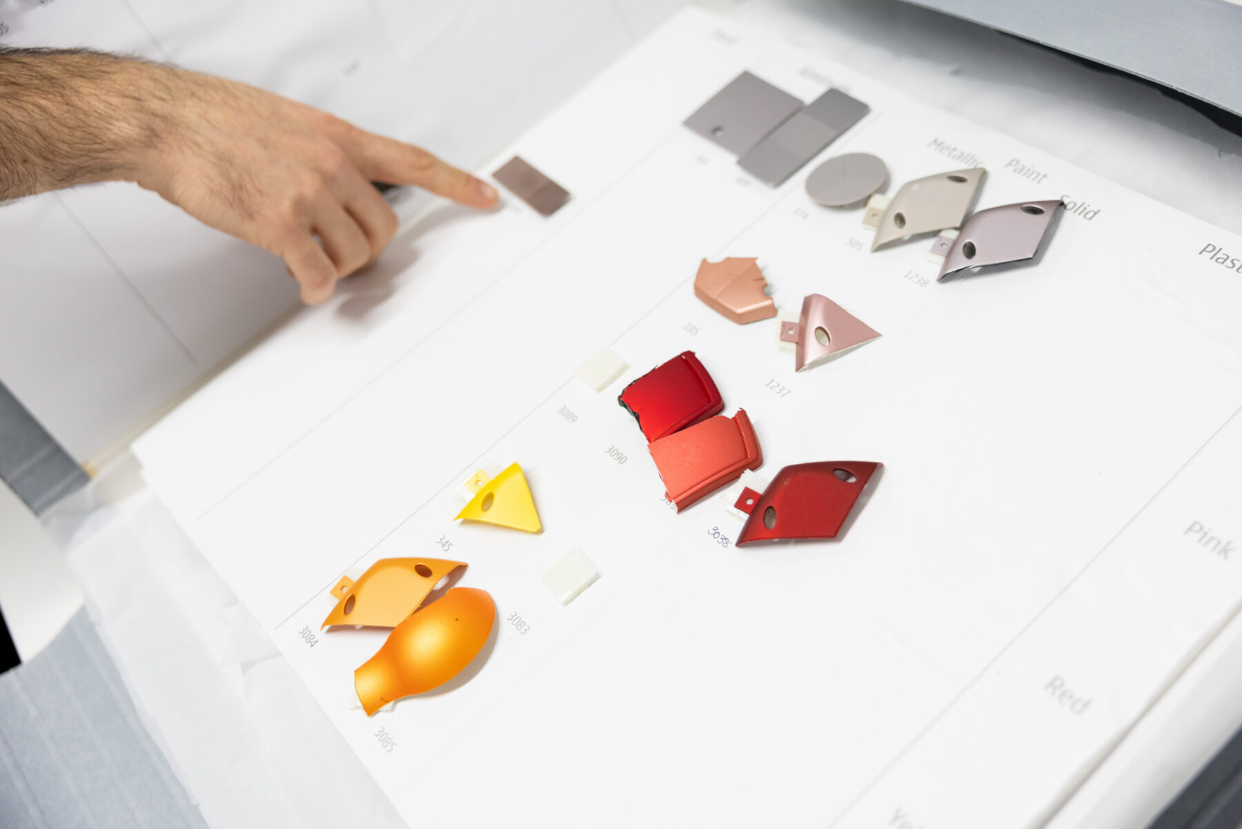 A hand points at a chart displaying an assortment of small coloured plastic and metal samples arranged in rows.