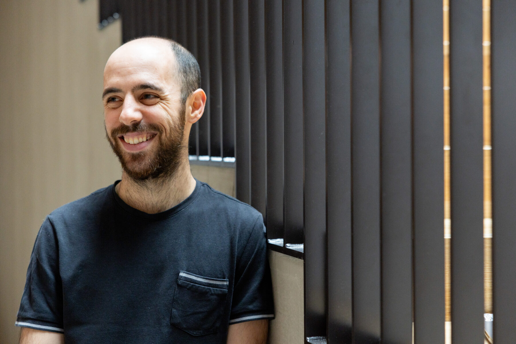 A man with a short beard and a dark T-shirt leans against a staircase wall beside tall vertical railing supports.