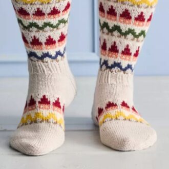A person stands wearing knee-high knitted socks decorated with colourful elf patterns on a light wooden floor.