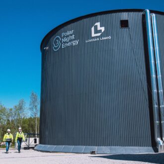 Duas pessoas caminham ao lado de um grande silo com tubos de metal descendo por sua lateral.