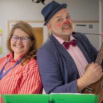 A woman in glasses smiles next to a man with clown makeup, a hat, a bowtie and a ukulele.