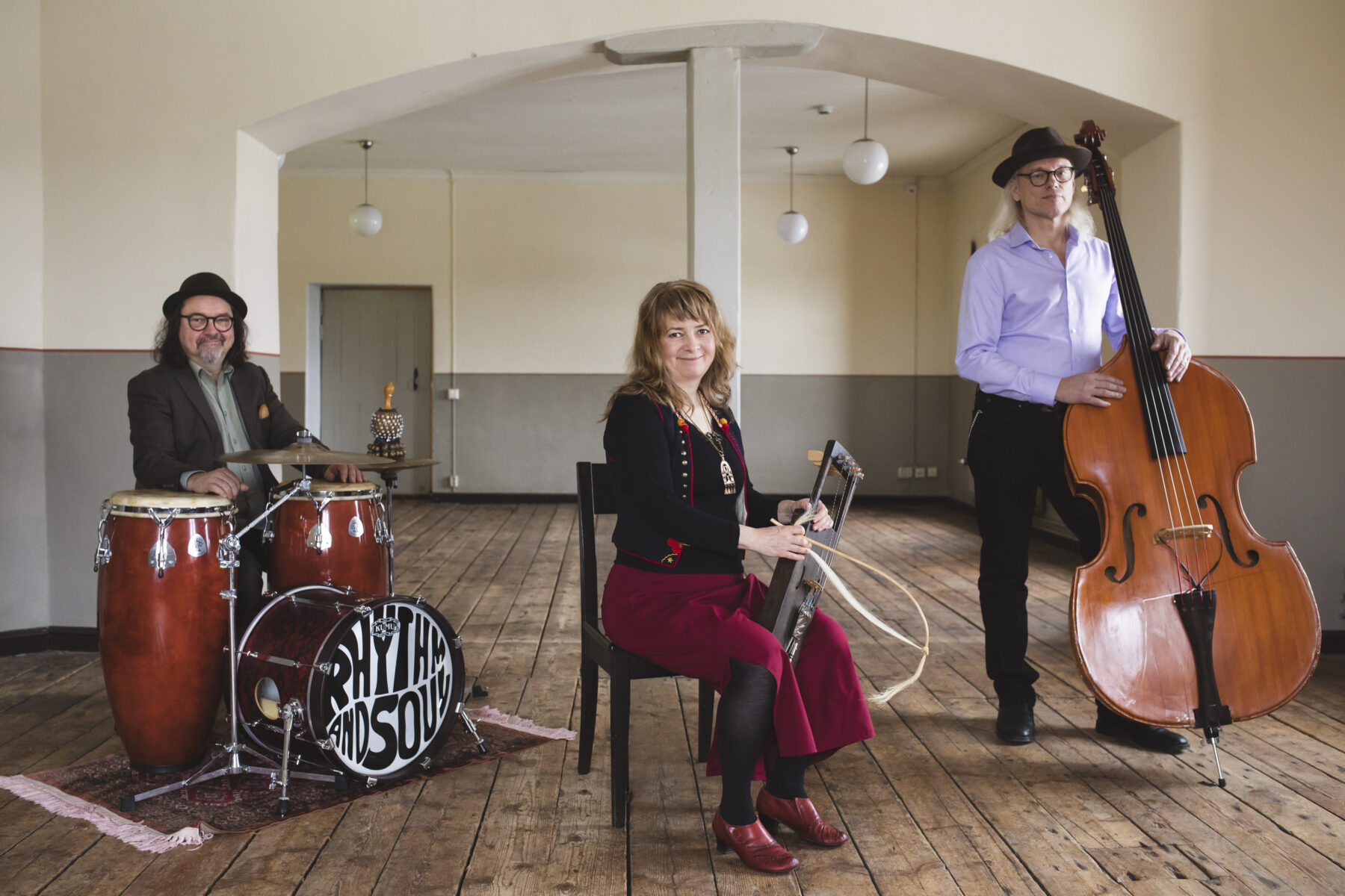 Three musicians are shown in a large room, with one sitting and holding a bowed lyre, one sitting in front of conga drums, and another standing beside a double bass.