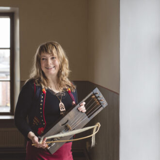 Tytti Metsä posa sonriente en el interior de una sala, junto a una ventana, mientras sostiene en sus manos un arpa de arco hecha de madera.