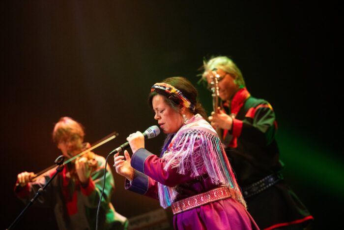 Ulla Pirttijärvi sings on stage with accompanying musicians under warm stage lighting.
