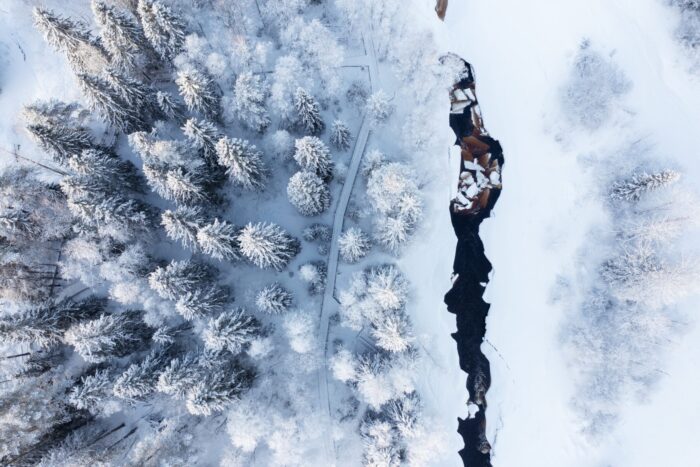 A snowy woodland seen from above is split by a narrow river flowing through the white landscape.