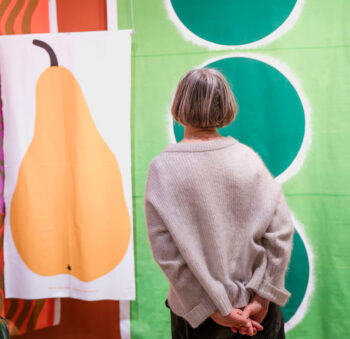 Eine Besucherin steht mit verschränkten Händen da und betrachtet Maija Isolas Textildruck mit dem Birnenmuster „Päärynä“.