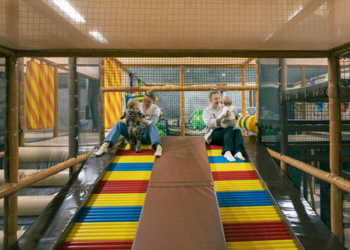  Two women hold their babies on a colourful cushioned slope inside a children’s play space.