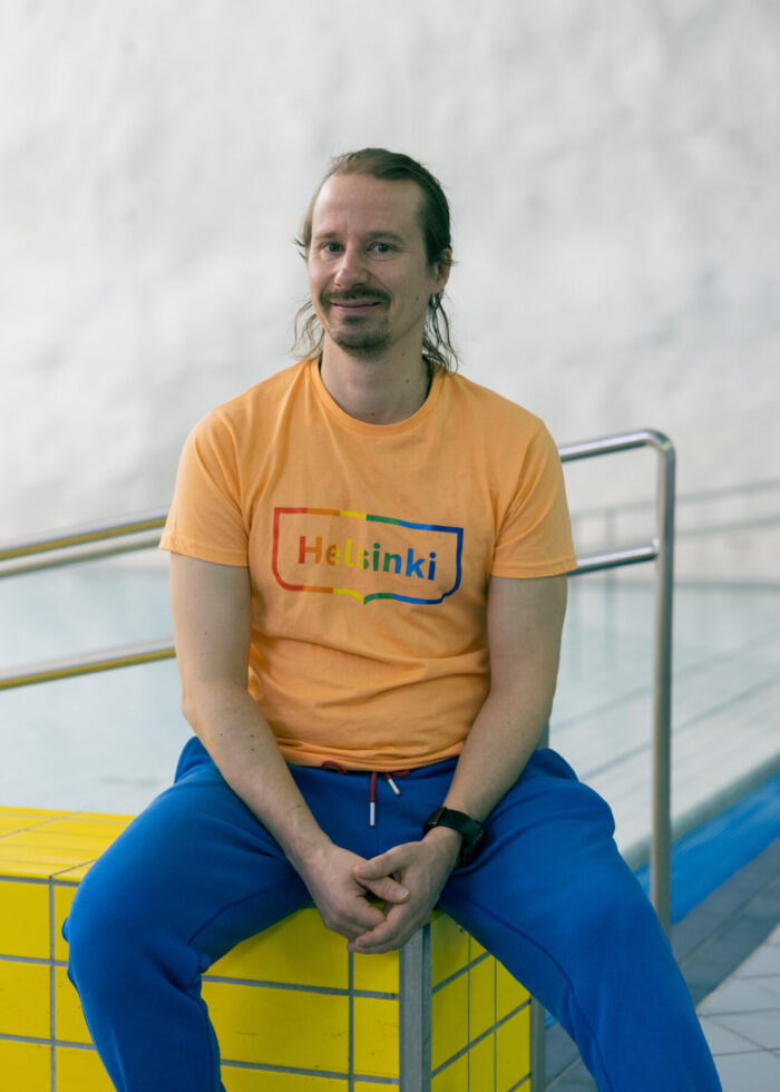 A man sits beside an indoor pool, dressed in a bright T-shirt and blue trousers.
