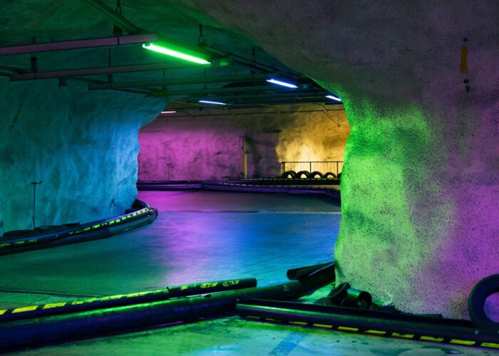 An empty underground karting track curves through a concrete tunnel lit with colourful neon lights and lined with tyre barriers.