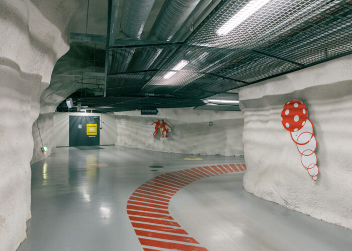 An empty underground shelter passage curves around rock-textured walls with smooth concrete floors and metal grating along the ceiling.
