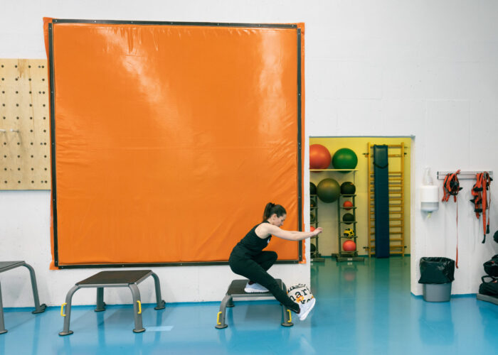 A woman performs a single-leg squat on a low bench in a brightly lit exercise room with blue flooring and a large orange wall mat.