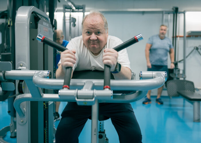 A man exercises on a gym machine, pulling handles towards him in a bright fitness space.
